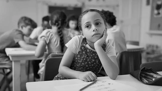Skolejente sitter ved pult i klasserom og ser tankefull ut mens andre elever arbeider i bakgrunnen.
