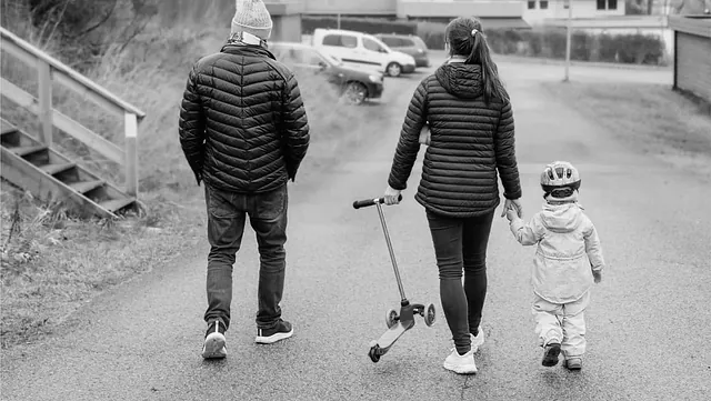 Familie med liten jente og sparkesykkel går langs en rolig boliggate i et nabolag.
